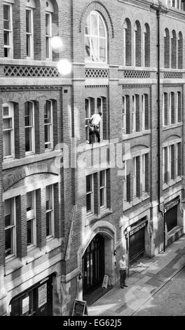 Window cleaner on very narrow ledge 3storeys up with no safety gear and man below oblivious to what is above him Stock Photo