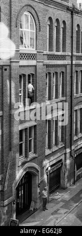 Window cleaner on very narrow ledge 3storeys up with no safety gear and man below oblivious to what is above him Stock Photo