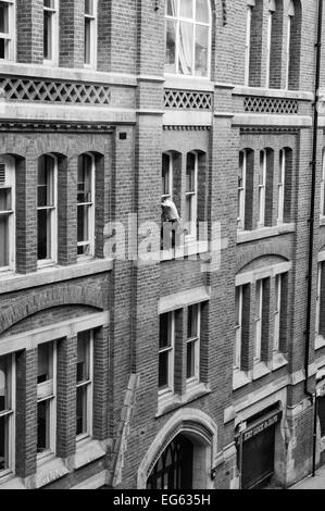 Window cleaner on very narrow ledge 3storeys up with no safety gear and man below oblivious to what is above him Stock Photo