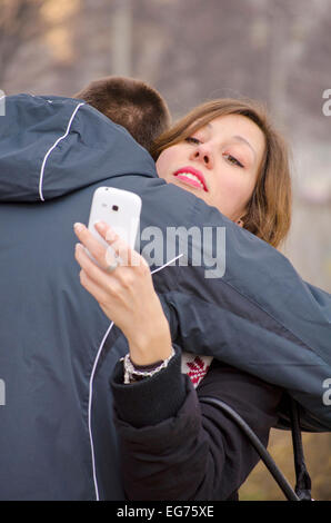 Boy hugging a girl while she looks at her smart phone outdoors Stock Photo