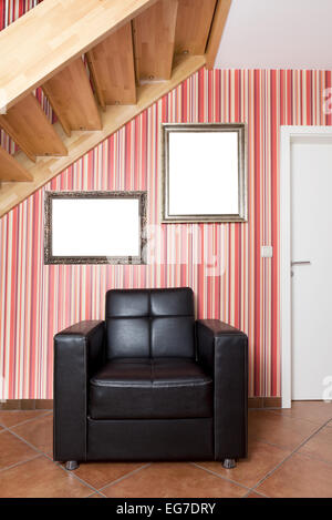 in a hallway with stairs is a leather armchair and empty frames hanging on the wall Stock Photo