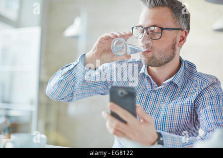 Businessman in casualwear drinking water while reading sms or dialing number on cellphone Stock Photo