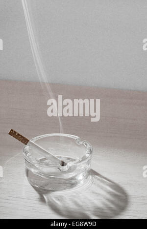A recently lid cigarette rests in a glass ashtray while the smoke floats up in the air. Stock Photo