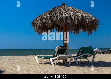 Comfortable and relaxing lounge chairs under a grassy palapa on a sandy beach on the ocean on a sunny day in Mexico. Stock Photo