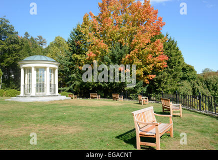 round gazebo and wood park benches by a black iron fence.  There is a grass area and trees, with leaves starting to change color Stock Photo