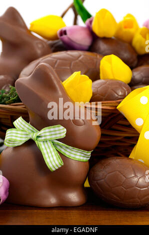 Happy Easter chocolate hamper of eggs and bunny rabbits in large basket with yellow and pink purple silk tulip flowers on dark w Stock Photo