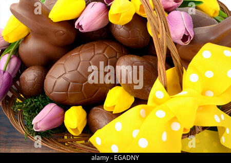 Happy Easter chocolate hamper of eggs and bunny rabbits in large basket with yellow and pink purple silk tulip flowers on dark w Stock Photo