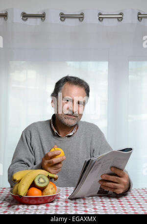 Middle aged man holding a magazine while eating fruit for breakfast in the kitchen at home Stock Photo