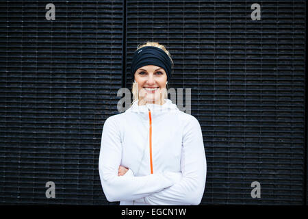 Portrait of happy young woman in sportswear looking at camera smiling. Fitness female against dark wall. Stock Photo