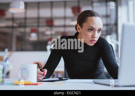 Image of young businesswoman looking at laptop while working at her desk. Female web designer taking notes from internet. Stock Photo
