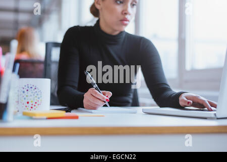 Young woman taking notes from laptop. Female executive working her desk using laptop and writing notes. Stock Photo