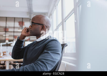 Side view of african executive sitting at his desk using mobile phone. Young man at work answering a phone call. Stock Photo