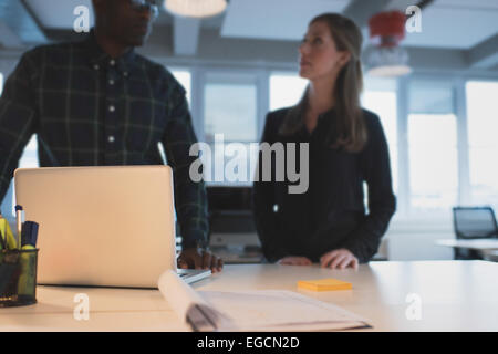 Table in office with business people standing by discussing. Focus on laptop and documents on table with colleagues around. Stock Photo