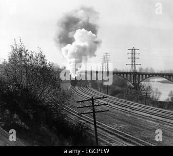 1940s AERIAL OF TRAIN TRAVELING ALONG RIVER UNDER BRIDGE BILLOWING SMOKE NEAR COLUMBUS OHIO Stock Photo