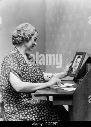 1930s 1940s SENIOR WOMAN SITTING AT DESK WRITING LETTER AS SHE HOLDS PICTURE PORTRAIT OF YOUNG MAN SON Stock Photo