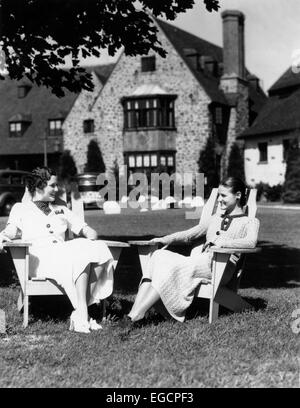 1930s TWO WOMEN SITTING IN ADIRONDACK LAWN CHAIRS IN FRONT OF STONE ESTATE HOUSE TALKING Stock Photo