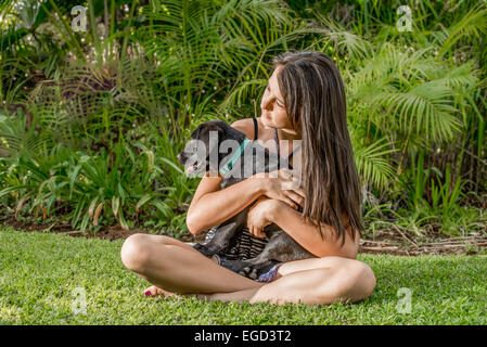 Young teenage girl sitting on lawn by the garden of her home while holding a black Labrador puppy in her arms and hands. Stock Photo