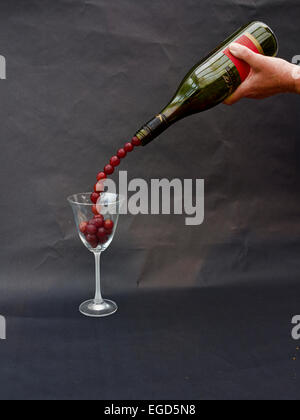 A bottle with red grapes falling into a wine glass giving the impression that wine is flowing into the glass Stock Photo