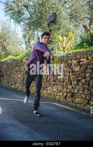 Angry young man standing in the middle of a rural road with a stone wall tossing his shoe through the air towards the camera Stock Photo