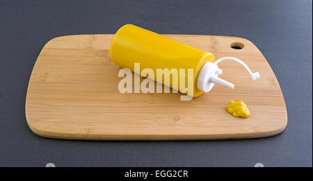 A bottle of mustard laying on its side with a small blob squeezed out in front onto a wood cutting board Stock Photo