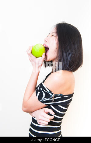 Young beautiful asian woman biting green apple with angry expression, profile on white background Stock Photo