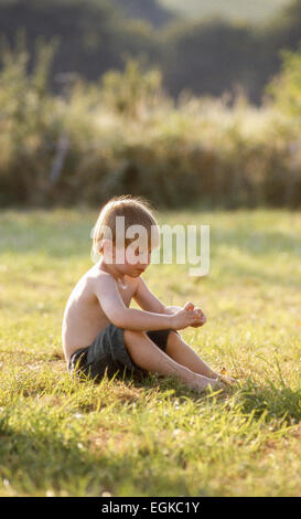 Young thoughtful barefoot boy sitting alone in sunny natural country green grass field with hedge in background Stock Photo
