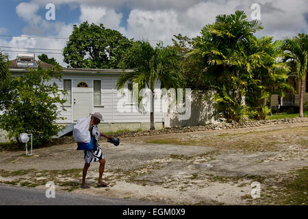 Barbados, white wooden house in background, trees and plants. Man walking, side view, carrying a white bag over shoulder Stock Photo