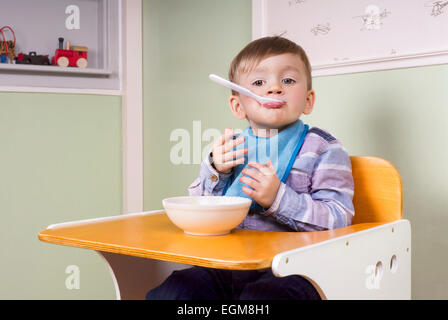 two year old boy sitting in his chair eating yoghurt and making fun Stock Photo