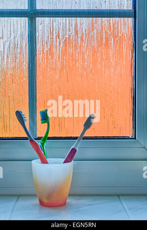 Orange plastic cup holding three toothbrushes (orange, green and pink) sitting on a bathroom windowsill against a frosted window Stock Photo