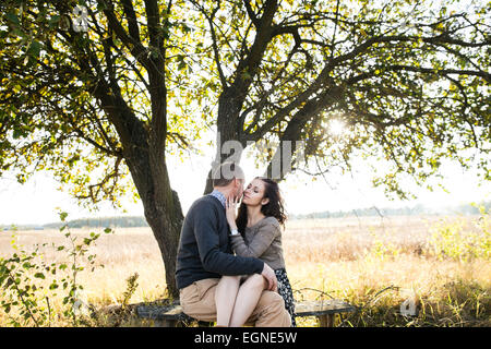 Image of a young couple kissing while sitting on a bench under tree at autumn sunset Stock Photo