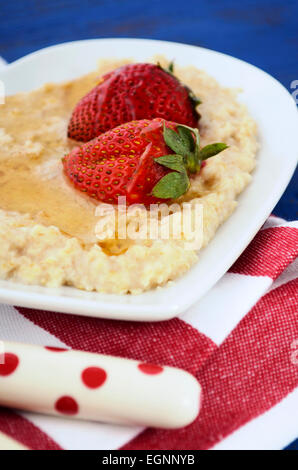 Plate of nutritious and healthy cooked breakfast oats with strawberries and honey in heart shaped bowl on dark blue rustic wood  Stock Photo