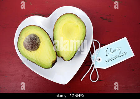 Avocado cut in half on heart shape plate on dark red vintage rustic wood table, with I Love Avocados text sign. Stock Photo