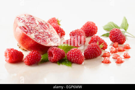 Raspberry, Rubus idaeus cultivar and Pomegranate, Punica cultivar cut in half surrounded with several raspberries and leaves on white marble. Selective focus. Stock Photo