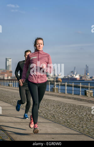 Fit active young couple running on a seafront promenade with the athletic woman leading the way approaching the camera in a heal Stock Photo