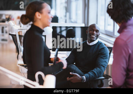 Happy african man sitting at desk with coworkers smiling. Young business executives during break. Stock Photo