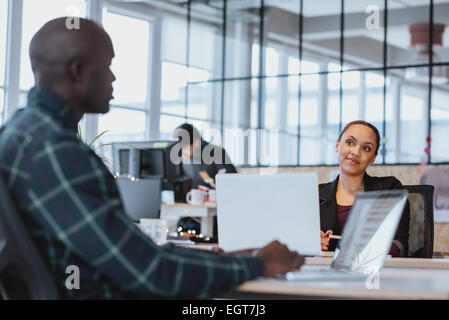 Young woman looking at male colleague while sitting at a table with laptop. African executive discussing work with coworker. Stock Photo