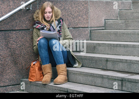 Trendy young woman in winter fashion sitting reading on outdoor, urban steps as she studies up her college notes or reads a jour Stock Photo