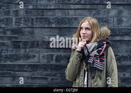 Thoughtful pretty blond girl standing in front of a textured grey brick wall with her hand to her chin staring pensively up into Stock Photo