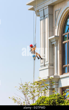 Window cleaner wearing orange overalls and red helmet, suspended from harness and ropes cleaning windows in Chennai, India Stock Photo