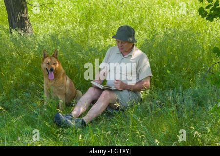 Mature man reading an interesting book to young dog under tree shadow Stock Photo