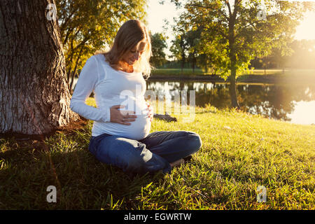 Pregnant woman sitting on the grass in a park at sunset Stock Photo