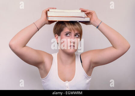 Close up Serious Young Woman with Short Blond Hair, Wearing Casual Sleeveless White Shirt, Balancing Books on her Head Stock Photo
