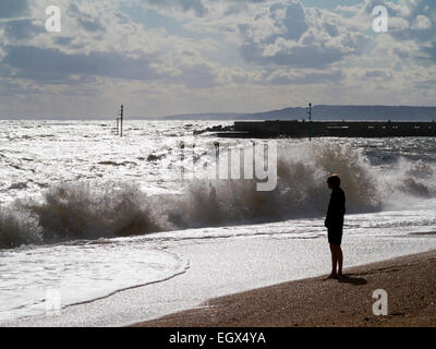 Teenage boy standing silhouetted on a beach looking at the sea with waves crashing on to the beach Stock Photo