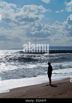 Teenage boy standing silhouetted on a beach looking at the sea with waves crashing on to the beach Stock Photo