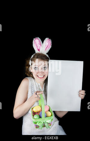 Young woman posing with rabbit ears, Easter basket with sequin eggs and blank sign for copy space on black background. Stock Photo