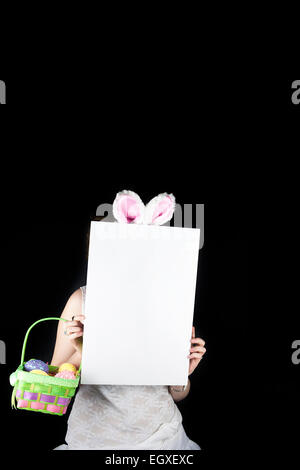 Young woman posing with rabbit ears, Easter basket with sequin eggs and blank sign for copy space on black background. Stock Photo