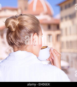 Back side on woman having morning coffee in the hotel, wearing white robe and enjoying cityscape from the window, Europe, Italy Stock Photo