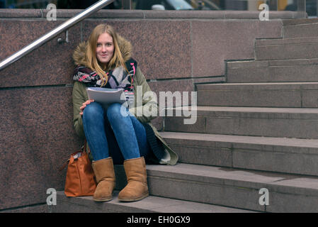 Trendy young woman in winter fashion sitting reading on outdoor, urban steps as she studies up her college notes or reading Stock Photo