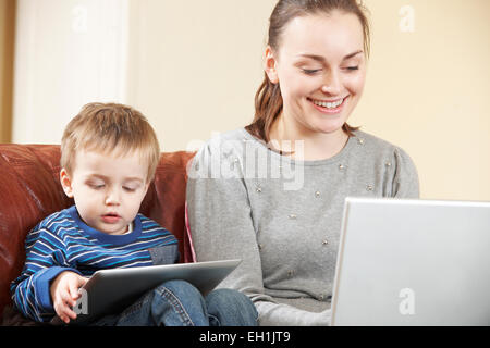 Mother Working On Laptop Whilst Son Uses Digital Tablet Stock Photo