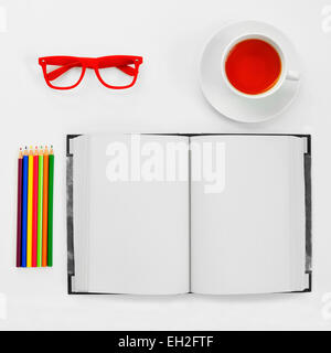 high-angle shot of a some colored pencils, a blank notebook, a pair of red plastic-rimmed eyeglasses and a cup of tea on a white Stock Photo